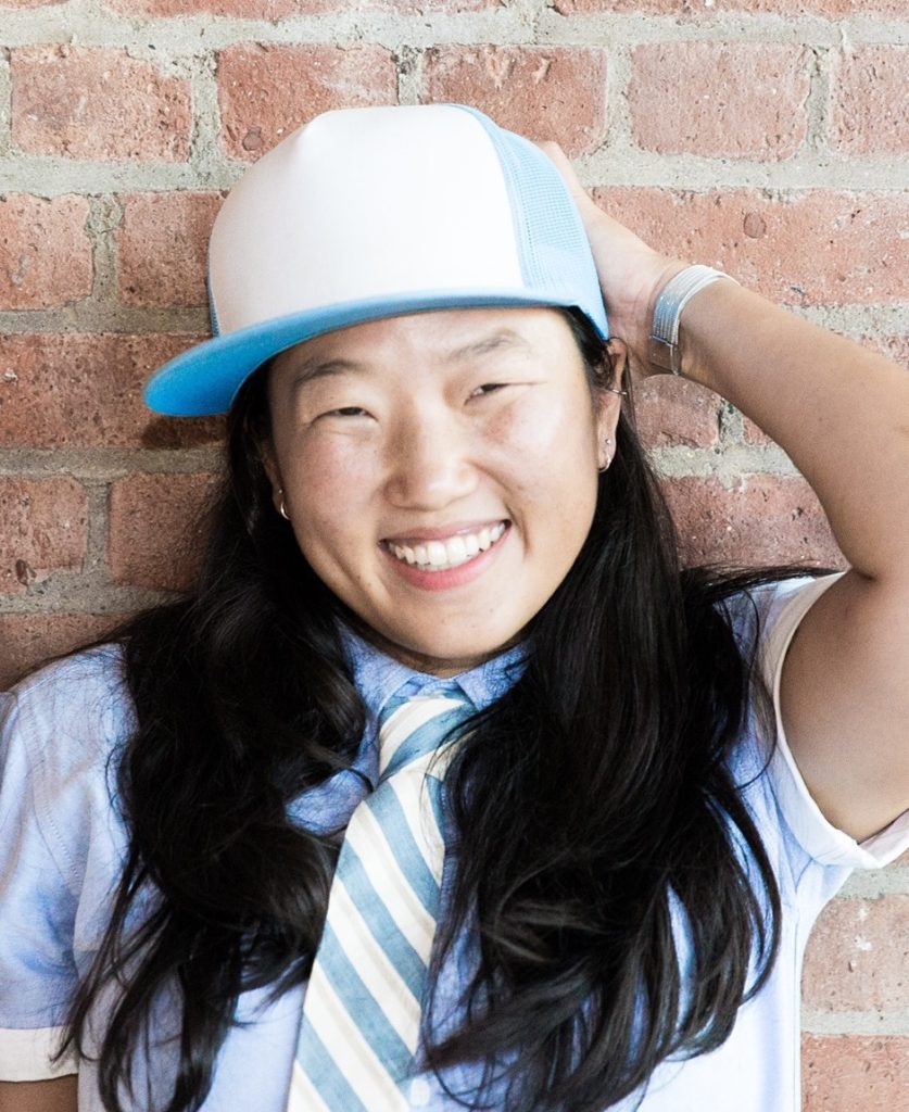 Photo of Arhm Choi Wild by Katharine Reece. Author with long, black hair, smiling broadly at the camera and wearing a baby blue-and-white baseball cap, a pale blue, short-sleeved,  buttoned shirt, rolled up at the sleeves, a watch with a white band, and a cream tie that has blue and taupe diagonal stripes. The subject's left arm is raised and her hand placed behind her head in a jaunty, carefree fashion.