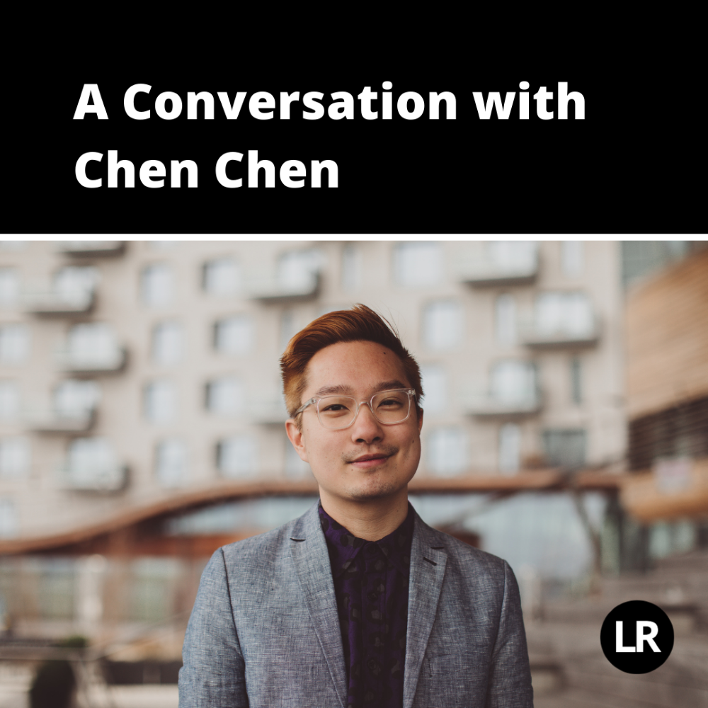 A Conversation with Chen Chen. Photo of Chen Chen, poet wearing clear glasses and with short brown hair, slicked back. He wears a dark shirt with a grey suit jacket and is standing against a blurred background of apartment buildings. 