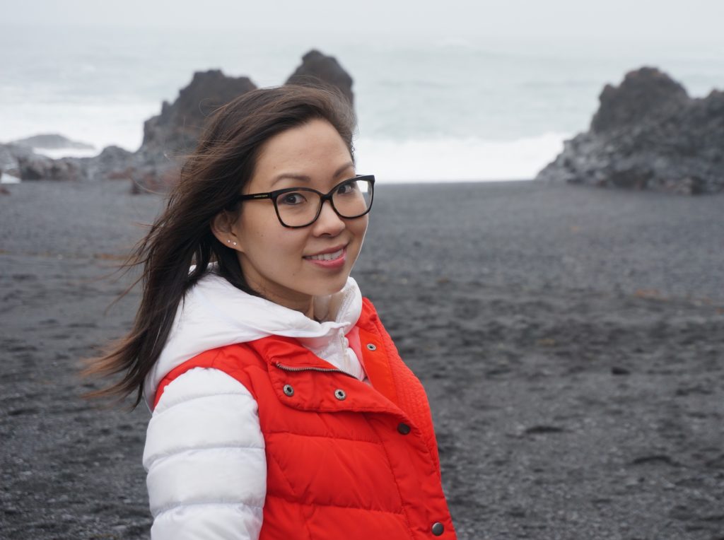 Photograph of Eugenia Leigh, poet with long, dark hair and thick-rimmed glasses. She is wearing a white, puffy jacket with a bright red vest layered on top and is standing in front of a moody seascape with rocky crags and crashing waves visible in the far distance.