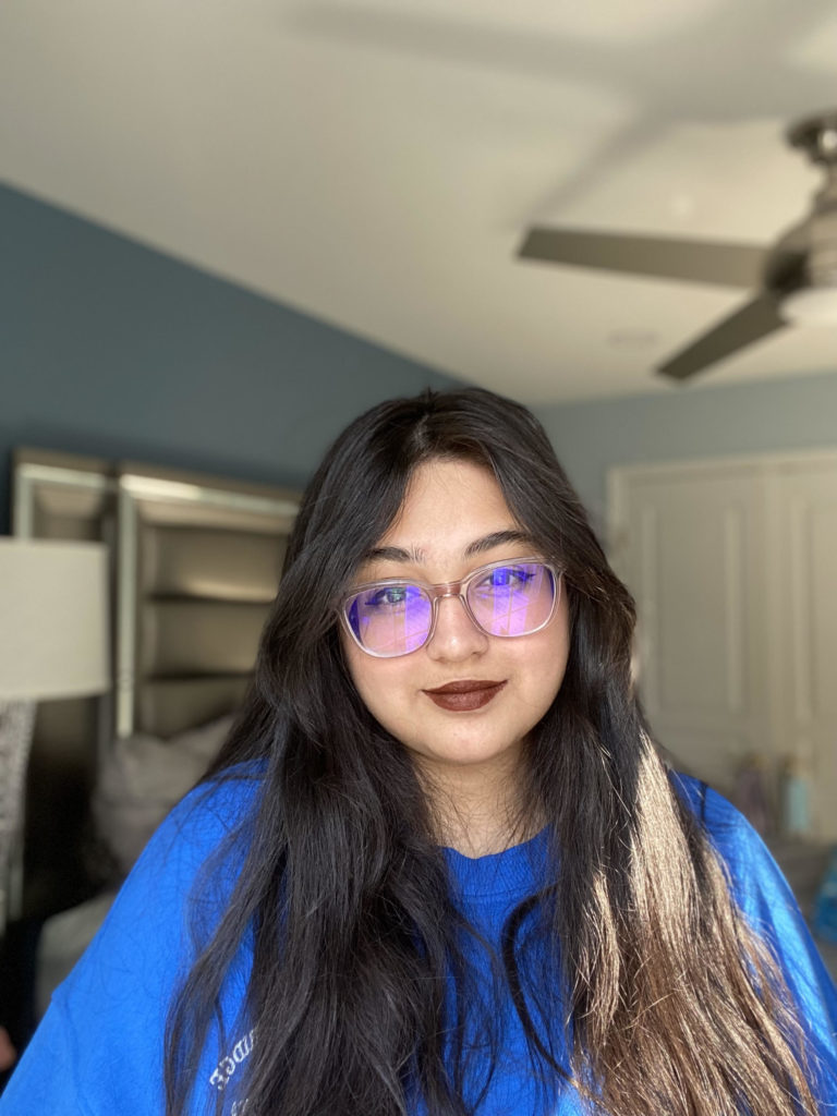 Close portrait of Pranaya S. Ayyala, Indian American poet, wearing a deep ocean blue sweatshirt and standing in front of a background of a gray bedroom. Her wavy brown hair is golden under the sunlight, falling a couple inches past her shoulders. She is wearing brown lipstick and large, light pink glasses. She is looking directly into the camera with a smile.