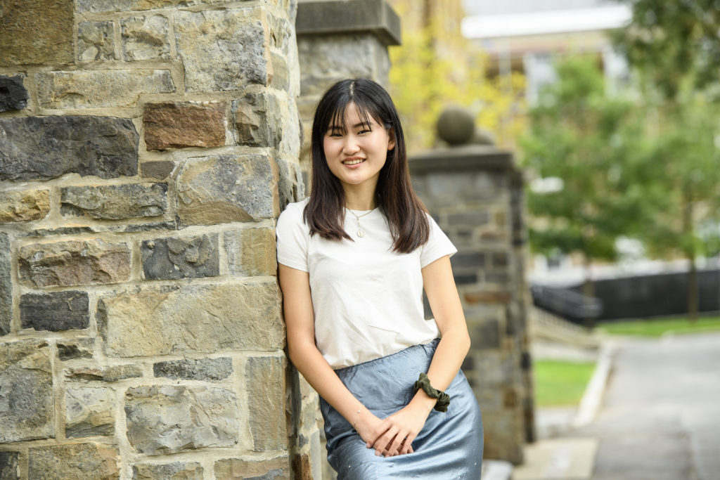 Photo of Rachel Lu, a Chinese American writer, scholar, and editor with mid-length black hair and bangs. She wears a white t-shirt, blue skirt, and gold necklace and looks into the camera while leaning against a stone wall with folded hands.