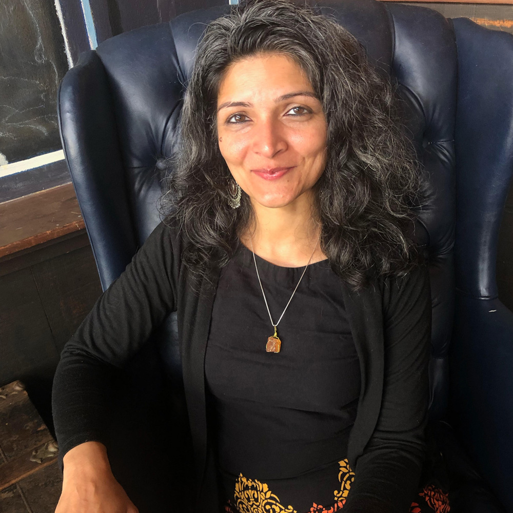 Portrait of Monica Mody, Indian American poet with shoulder-length, wavy, dark hair. She wears a black top and cardigan; a black skirt with an ornate yellow and orange print; large, lacy pendant earrings; and an amber-colored stone on a silver necklace. She is sitting in a leathery blue wingback arm chair and smiling slightly at the camera.